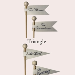 Puede incluir: Cuatro banderas de tela color hueso unidas a palos de madera con remates redondos. Dos banderas son rectangulares con 'The Vances' y 'The Tannenbaums' impresos en negro. Dos son triangulares con 'The Sims' y 'The Gutierrez' impresos en negro.