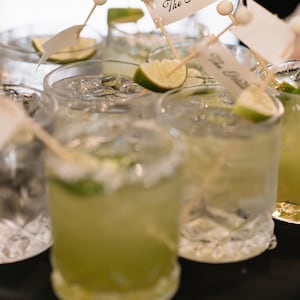 Puede incluir: Varios vasos transparentes llenos de un l&iacute;quido verde p&aacute;lido, cubitos de hielo y rodajas de lima. Cada vaso contiene un palillo de c&oacute;ctel de madera con una cuenta blanca y una peque&ntilde;a bandera de papel blanco con el texto The Hills impreso.