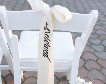 May include: A white folding chair featuring a long, cream-colored satin ribbon tied in a bow around the top backrest. The ribbon hangs vertically and displays the word Reserved printed in a black, elegant, cursive script font.