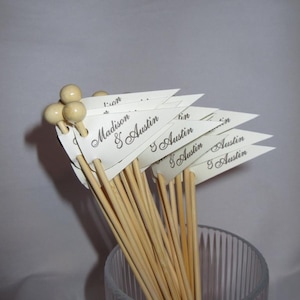 Puede incluir: Un conjunto de brochetas de madera rematadas con peque&ntilde;as cuentas de madera redondas. Cada brocheta presenta una bandera triangular de papel blanco impresa con los nombres Madison & Austin en una fuente cursiva oscura. Las brochetas est&aacute;n en un vaso.
