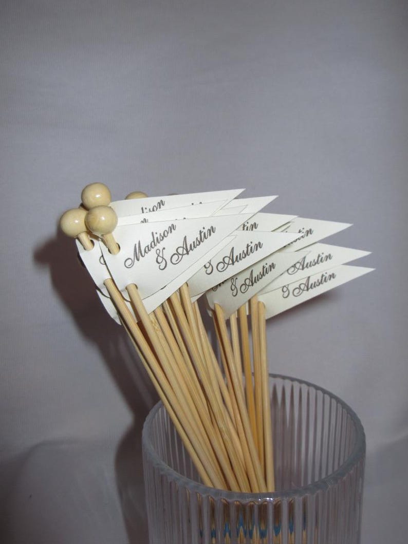 Puede incluir: Un conjunto de brochetas de madera rematadas con peque&ntilde;as cuentas de madera redondas. Cada brocheta presenta una bandera triangular de papel blanco impresa con los nombres Madison & Austin en una fuente cursiva oscura. Las brochetas est&aacute;n en un vaso.