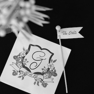 Puede incluir: Una servilleta cuadrada blanca con un escudo negro impreso que presenta una letra G central, detalles florales y la fecha 10.11.25 en una cinta. Un peque&ntilde;o palillo en forma de bandera blanca con el texto The Hills se coloca junto a la servilleta.