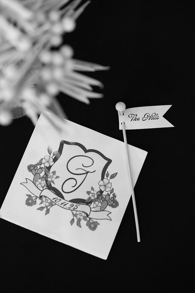Puede incluir: Una servilleta cuadrada blanca con un escudo negro impreso que presenta una letra G central, detalles florales y la fecha 10.11.25 en una cinta. Un peque&ntilde;o palillo en forma de bandera blanca con el texto The Hills se coloca junto a la servilleta.