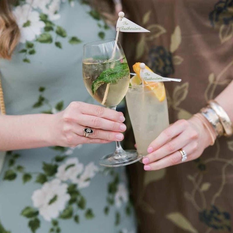 Puede incluir: Dos recipientes de cristal transparente con bebidas claras, cada uno adornado con un peque&ntilde;o palillo de bandera triangular blanca con la etiqueta The Featherstons. Un vaso contiene una guarnici&oacute;n de hierbas verdes y el otro una rodaja de c&iacute;trico.