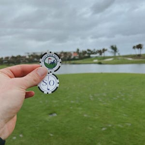 May include: Two round white golf ball markers with black edges. The top marker features a green golf course graphic with a palm tree, a red flag, and the text "WEST PALM BEACH 2025". The bottom marker displays the letters "SO" with a golf club icon.