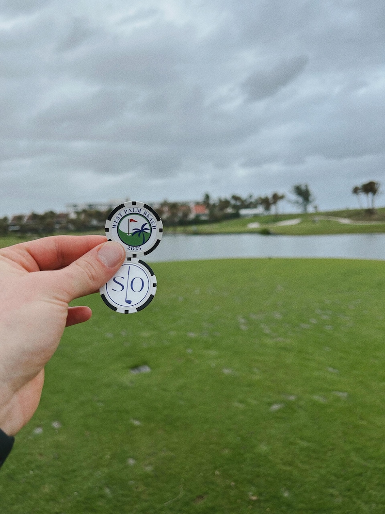 May include: Two round white golf ball markers with black edges. The top marker features a green golf course graphic with a palm tree, a red flag, and the text "WEST PALM BEACH 2025". The bottom marker displays the letters "SO" with a golf club icon.