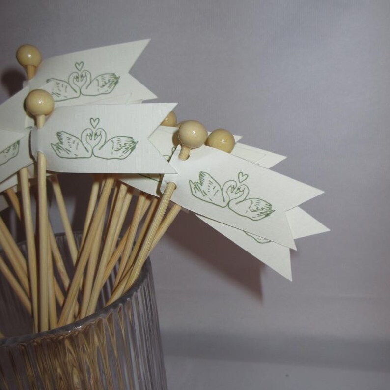 Puede incluir: Un conjunto de palillos de madera con peque&ntilde;as banderas de papel color crema en la parte superior. Cada bandera presenta una ilustraci&oacute;n en tinta verde de dos cisnes frente a frente con un peque&ntilde;o coraz&oacute;n entre sus cabezas.
