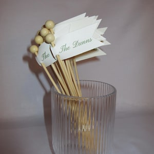 Puede incluir: Un conjunto de brochetas de madera con extremos esf&eacute;ricos de color claro y banderas de papel blanco. Cada bandera presenta el texto The Dunns impreso en una fuente cursiva verde. Las brochetas est&aacute;n agrupadas dentro de un vaso de vidrio estriado.