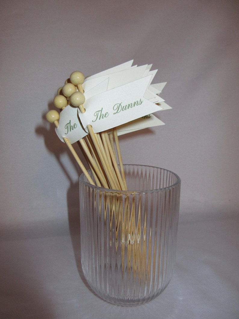 Puede incluir: Un conjunto de brochetas de madera con extremos esf&eacute;ricos de color claro y banderas de papel blanco. Cada bandera presenta el texto The Dunns impreso en una fuente cursiva verde. Las brochetas est&aacute;n agrupadas dentro de un vaso de vidrio estriado.