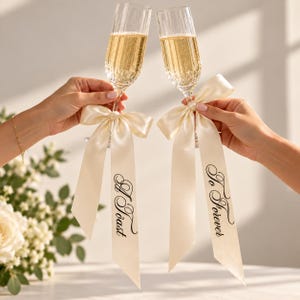 Puede incluir: Dos copas de champán transparentes y estriadas llenas de líquido dorado espumoso. Cada copa está adornada con una cinta de satén color crema atada en un lazo. Las cintas tienen el texto impreso en negro A Toast y To Forever.
