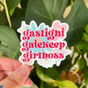 Peut inclure: Un autocollant avec un motif floral rose, bleu et blanc. L'autocollant dit "gaslight gatekeep girlboss" en lettres roses.