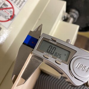 May include: A silver digital caliper measuring a piece of wood. The caliper reads 0.0825 inches.