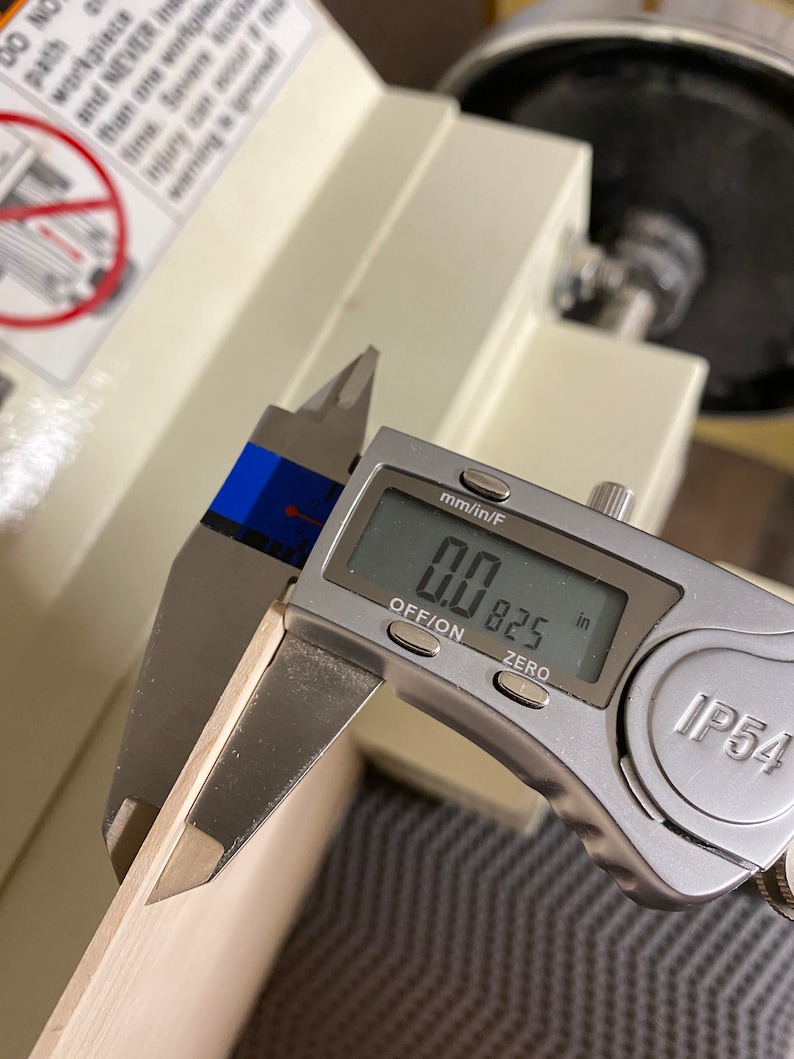 May include: A silver digital caliper measuring a piece of wood. The caliper reads 0.0825 inches.