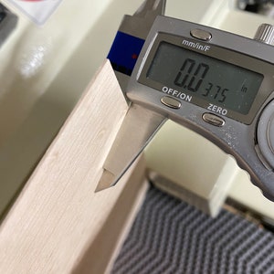 May include: A silver digital caliper measuring 0.375 inches. The caliper is measuring a piece of light wood.