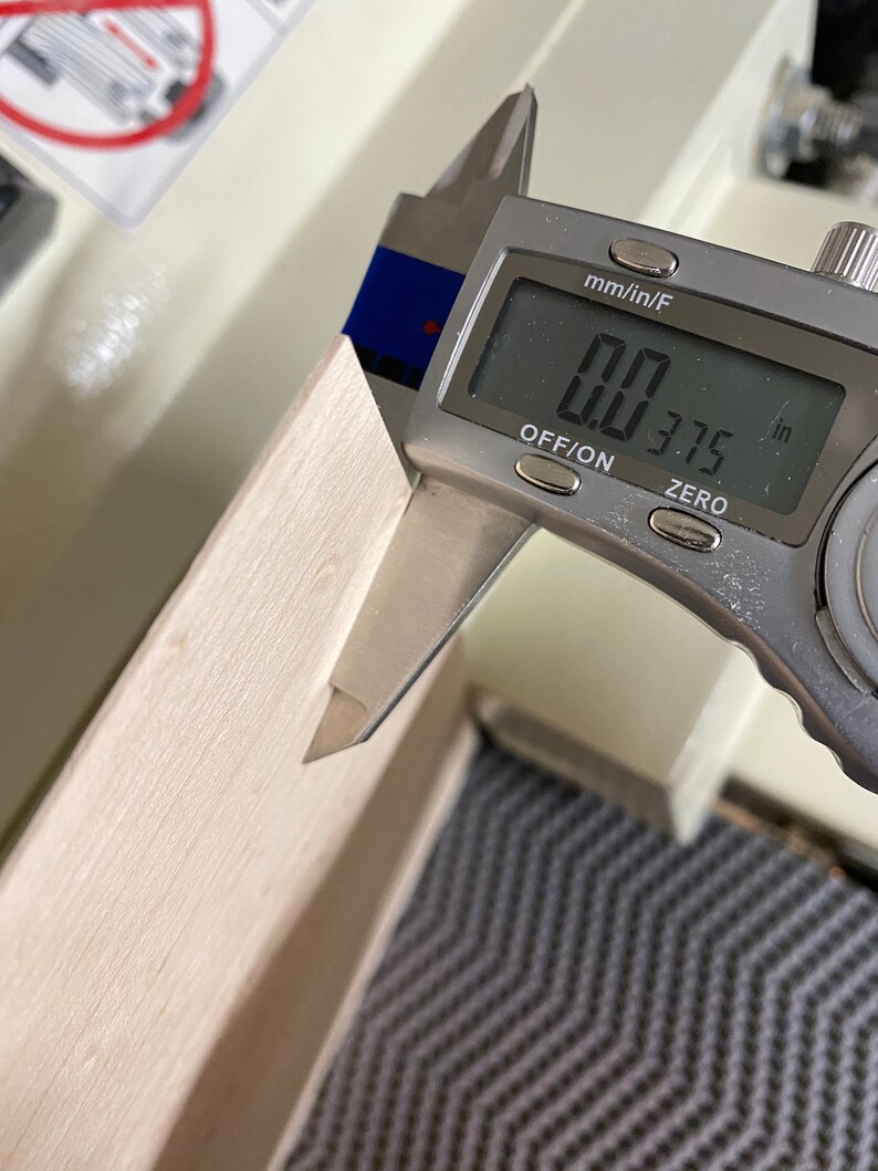 May include: A silver digital caliper measuring 0.375 inches. The caliper is measuring a piece of light wood.