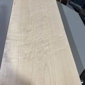 May include: A close-up of a light wood board with a natural wood grain pattern. The wood has a light brown color and a smooth surface.