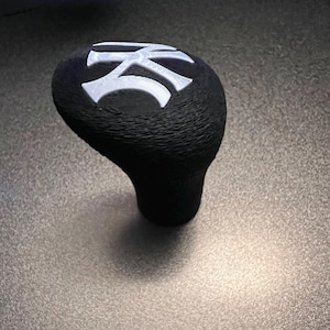 Puede incluir: Pomo de cambio negro con un diseño estilizado blanco en la parte superior.