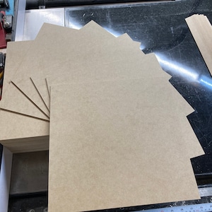 May include: A stack of eight light brown wood panels, each measuring approximately 12 inches by 12 inches. The panels are arranged in a staggered pattern, with the top panel slightly offset from the one below it. The panels are sitting on a black surface.