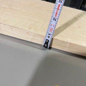May include: A white tape measure with black markings is measuring a piece of light wood. The tape measure reads "CRAFTSMAN" and has markings for inches and feet.
