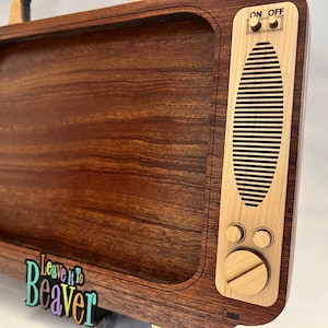 May include: A wooden TV tray with a brown wood finish and a light wood trim. The tray has a cutout in the shape of a television screen and features the text "Leave it to Beaver" in colorful letters. The tray has a small knob and two buttons on the side, labeled "ON OFF".