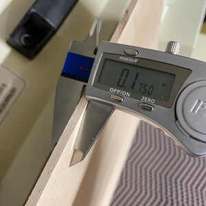 May include: A silver digital caliper measuring 0.1750 inches. The caliper is measuring the thickness of a piece of wood.