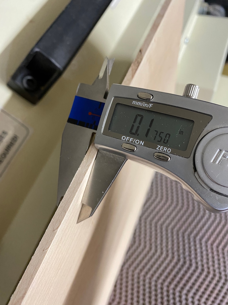 May include: A silver digital caliper measuring 0.1750 inches. The caliper is measuring the thickness of a piece of wood.
