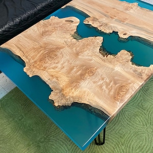 May include: A handcrafted coffee table with a unique design. The table features a light-colored wood top with a teal-colored epoxy resin river. The table is supported by black metal hairpin legs. The table measures approximately 48 inches long and 24 inches wide.