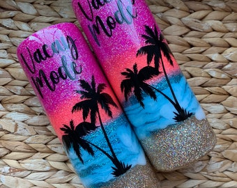 May include: Two pink and blue glitter tumblers with a sunset and palm tree design. The tumblers have the text "Vacation Mode" on them.