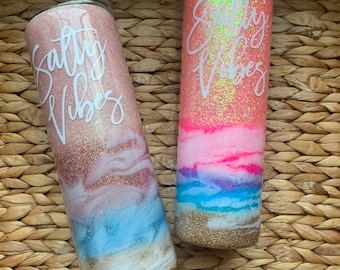 Könnte beinhalten: Zwei rosa und goldene Glitzerbecher mit dem Text "Salty Vibes" in Weiß. Die Becher haben ein Strandmotiv mit blauen und weißen Wellen und goldenem Sand.