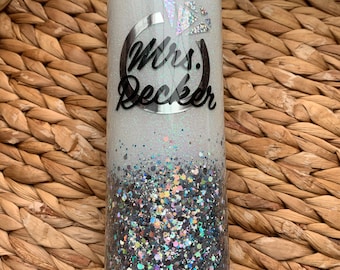 May include: White and silver glitter tumbler with a silver ring and the text "Mrs. Decker" in black lettering.