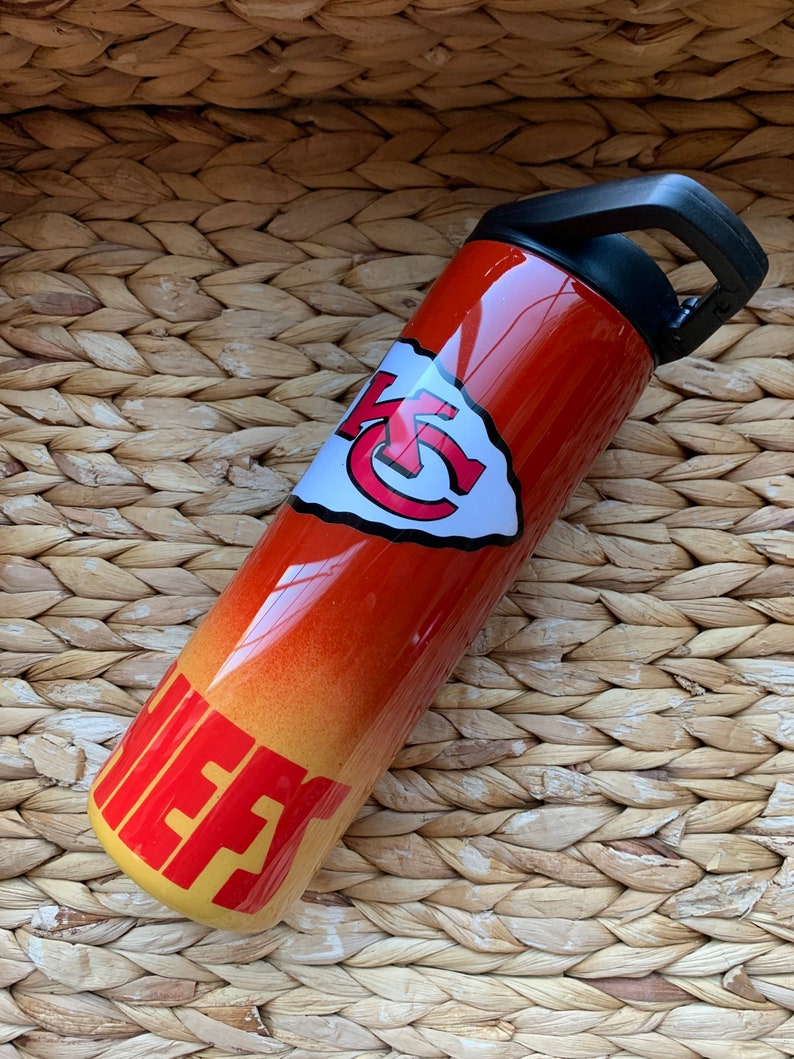 May include: A red and yellow stainless steel water bottle with a Kansas City Chiefs logo and the word "Chiefs" on the side. The bottle has a black lid with a carrying handle.