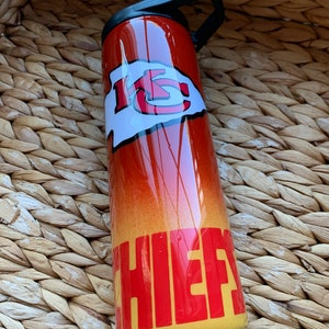 May include: A red and yellow stainless steel tumbler with a black lid and handle. The tumbler has a white and red "KC" logo and the word "CHIEFS" in red letters.