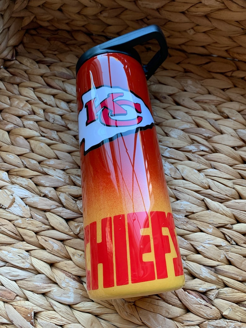 May include: A red and yellow stainless steel tumbler with a black lid and handle. The tumbler has a white and red "KC" logo and the word "CHIEFS" in red letters.