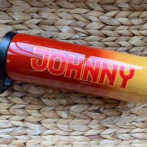 May include: A red and yellow ombre stainless steel tumbler with a black lid and the name "JOHNNY" printed in red on the side.