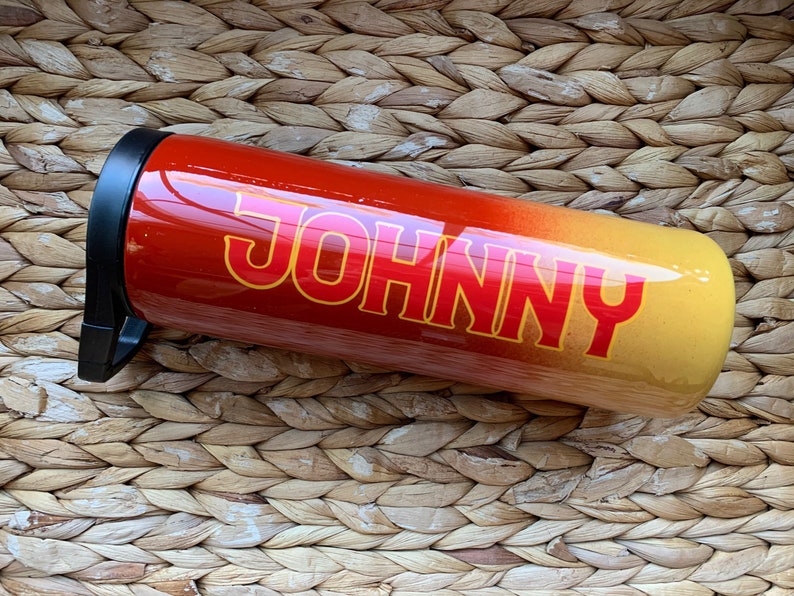 May include: A red and yellow ombre stainless steel tumbler with a black lid and the name "JOHNNY" printed in red on the side.