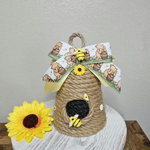 May include: A brown and tan beehive-shaped doorstop with a yellow sunflower and a Winnie the Pooh-themed bow. The doorstop is made of twine and has a small opening in the front.