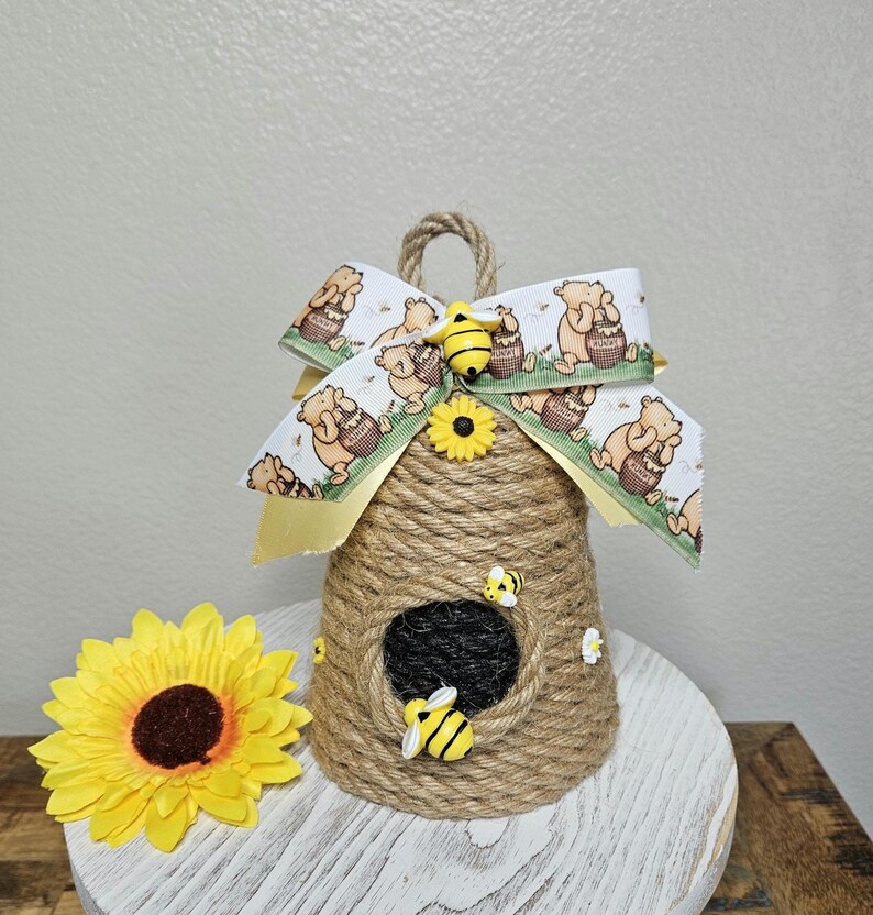 May include: A brown and tan beehive-shaped doorstop with a yellow sunflower and a Winnie the Pooh-themed bow. The doorstop is made of twine and has a small opening in the front.