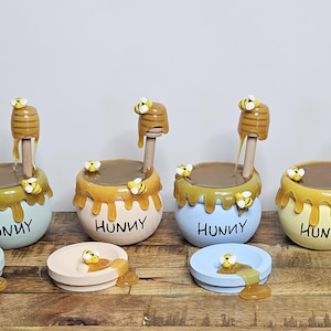 May include: A set of four ceramic honey pots in pastel colors: light blue, peach, and yellow. Each pot has a honey dipper and is decorated with bees and dripping honey. The word "HUNNY" is written on the front.