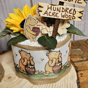 May include: A Winnie the Pooh themed diaper cake with a wooden sign that says "Welcome to the Hundred Acre Wood". The cake is decorated with sunflowers, Winnie the Pooh characters, and a honey pot.