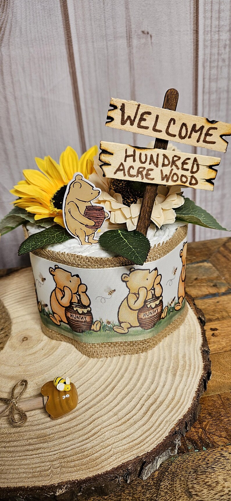 May include: A Winnie the Pooh themed diaper cake with a wooden sign that says "Welcome to the Hundred Acre Wood". The cake is decorated with sunflowers, Winnie the Pooh characters, and a honey pot.