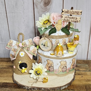 May include: A Winnie the Pooh themed baby shower diaper cake with a wooden sign that says "Welcome to the Hundred Acre Wood". The cake is decorated with Winnie the Pooh characters, honey pots, and bees. There is also a small beehive-shaped decoration with a bee on it.