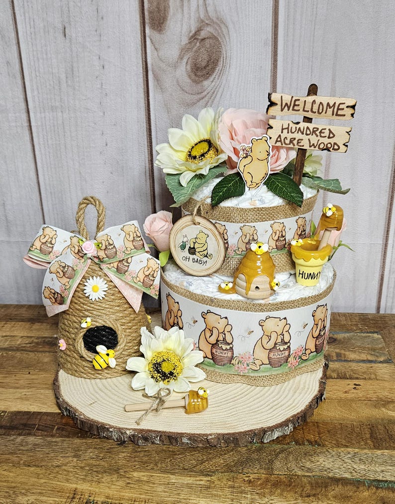 May include: A Winnie the Pooh themed baby shower diaper cake with a wooden sign that says "Welcome to the Hundred Acre Wood". The cake is decorated with Winnie the Pooh characters, honey pots, and bees. There is also a small beehive-shaped decoration with a bee on it.