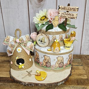 May include: A Winnie the Pooh themed diaper cake with a wooden sign that says "Welcome to the Hundred Acre Wood". The cake is decorated with Winnie the Pooh characters, honey pots, and bees. There is also a small beehive made of twine and a wooden honey dipper.