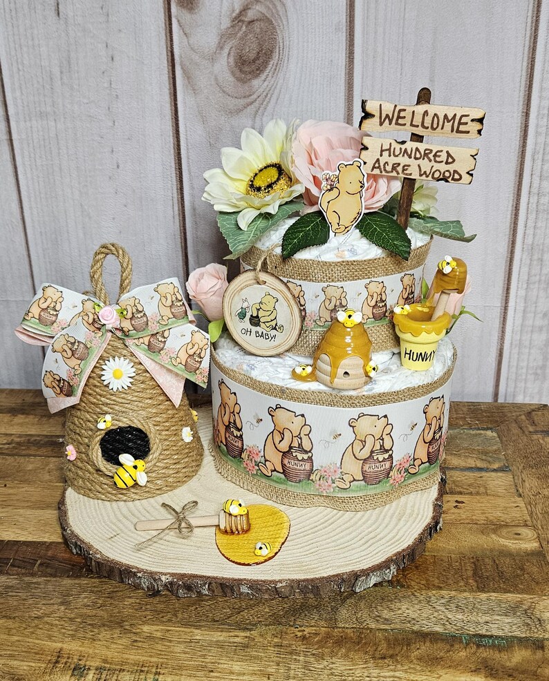 May include: A Winnie the Pooh themed diaper cake with a wooden sign that says "Welcome to the Hundred Acre Wood". The cake is decorated with Winnie the Pooh characters, honey pots, and bees. There is also a small beehive made of twine and a wooden honey dipper.