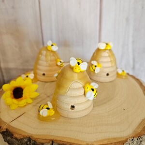 May include: Three wooden beehive-shaped ornaments with honey dripping down the sides. Each beehive is decorated with small yellow and black bees. The beehives are sitting on a wooden slice.
