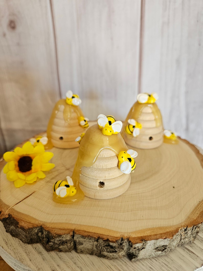 May include: Three wooden beehive-shaped ornaments with honey dripping down the sides. Each beehive is decorated with small yellow and black bees. The beehives are sitting on a wooden slice.