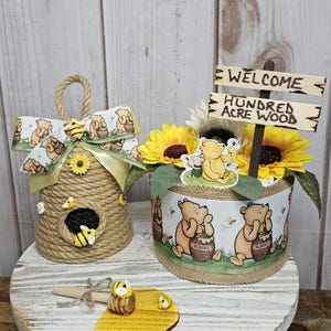 May include: A Winnie the Pooh themed baby shower centerpiece with a welcome sign that reads "Welcome Hundred Acre Wood". The centerpiece features a brown and yellow sunflower arrangement, a Winnie the Pooh character, and a brown and yellow honey pot. The centerpiece is on a white wooden table with a brown and yellow honey dipper and a honey comb.