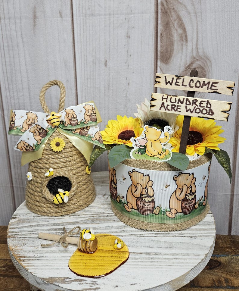 May include: A Winnie the Pooh themed baby shower centerpiece with a welcome sign that reads "Welcome Hundred Acre Wood". The centerpiece features a brown and yellow sunflower arrangement, a Winnie the Pooh character, and a brown and yellow honey pot. The centerpiece is on a white wooden table with a brown and yellow honey dipper and a honey comb.
