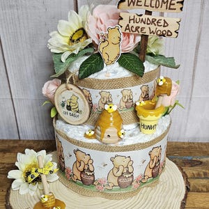 May include: A two-tiered Winnie the Pooh diaper cake decorated with Winnie the Pooh figurines, honey pots, and bees. The cake is topped with a wooden sign that reads "Welcome to the Hundred Acre Wood".