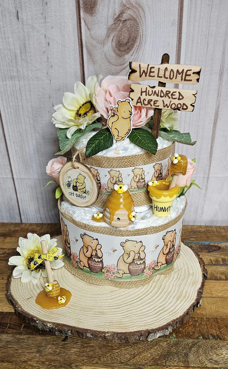 May include: A two-tiered Winnie the Pooh diaper cake decorated with Winnie the Pooh figurines, honey pots, and bees. The cake is topped with a wooden sign that reads "Welcome to the Hundred Acre Wood".